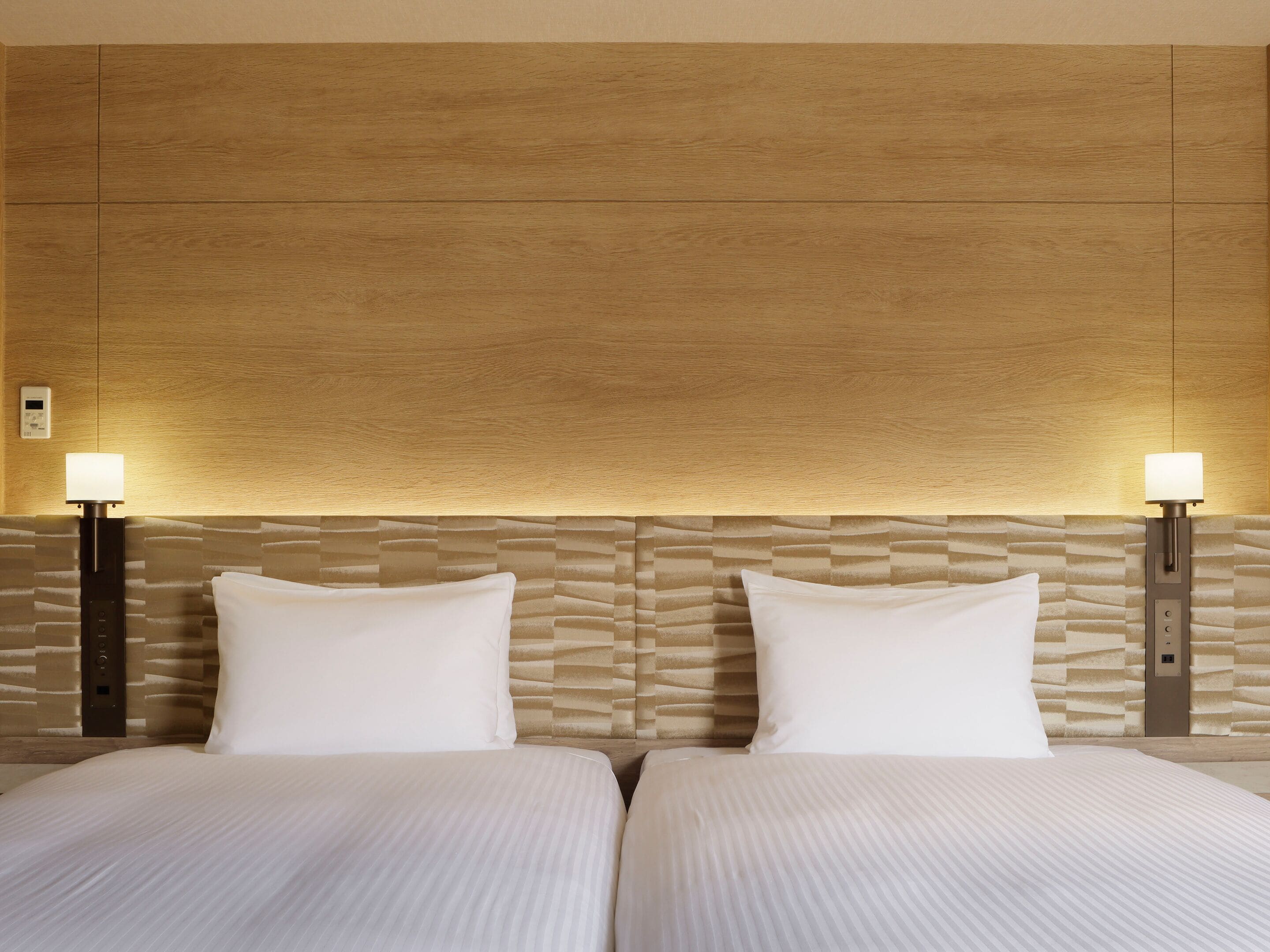 A hotel room with two beds, a textured headboard, and wall-mounted lamps.