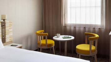 A hotel room with a bed, two yellow chairs, a small table with a cup and glasses, and a window with curtains.