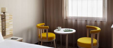 A hotel room with a bed, two yellow chairs, a small table with a cup and glasses, and a window with curtains.
