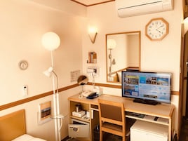 Habitación de hotel con escritorio, silla, computadora, mesita de noche, lámpara, reloj y una caja de seguridad pequeña.