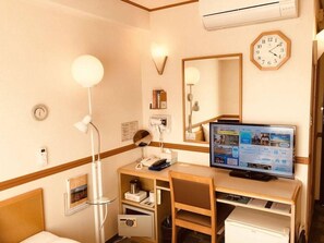 A hotel room with a desk, chair, computer, bedside table, lamp, and a clock on the wall.