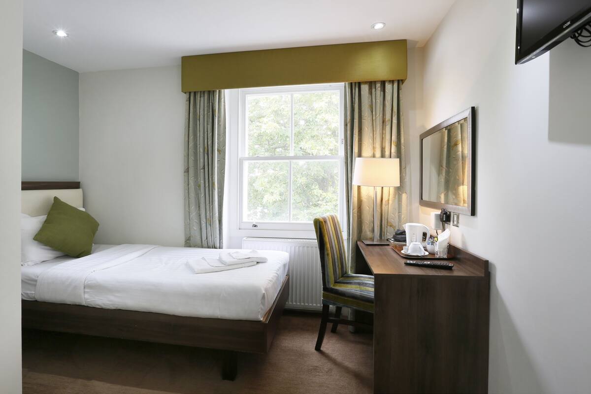 Una habitación de hotel con cama, escritorio, silla, televisor y ventana con cortinas.