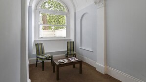 Une pièce dotée d’une grande fenêtre en arc de cercle, de deux fauteuils à rayures, d’une petite table avec des magazines et d’un mur blanc orné de moulures décoratives.