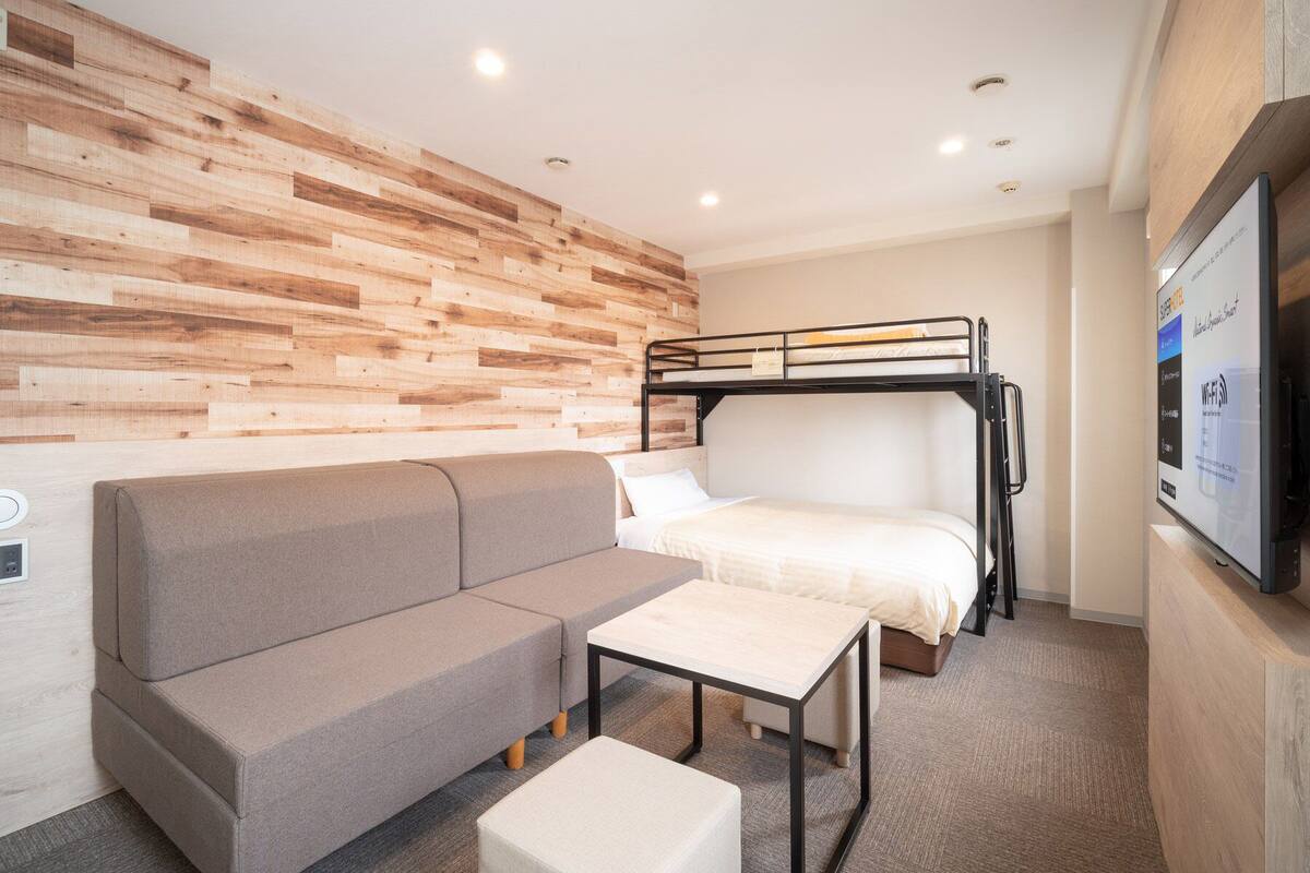 Una habitación de hotel moderna con una cama litera, un sofá, una mesita pequeña y un televisor montado en la pared.
