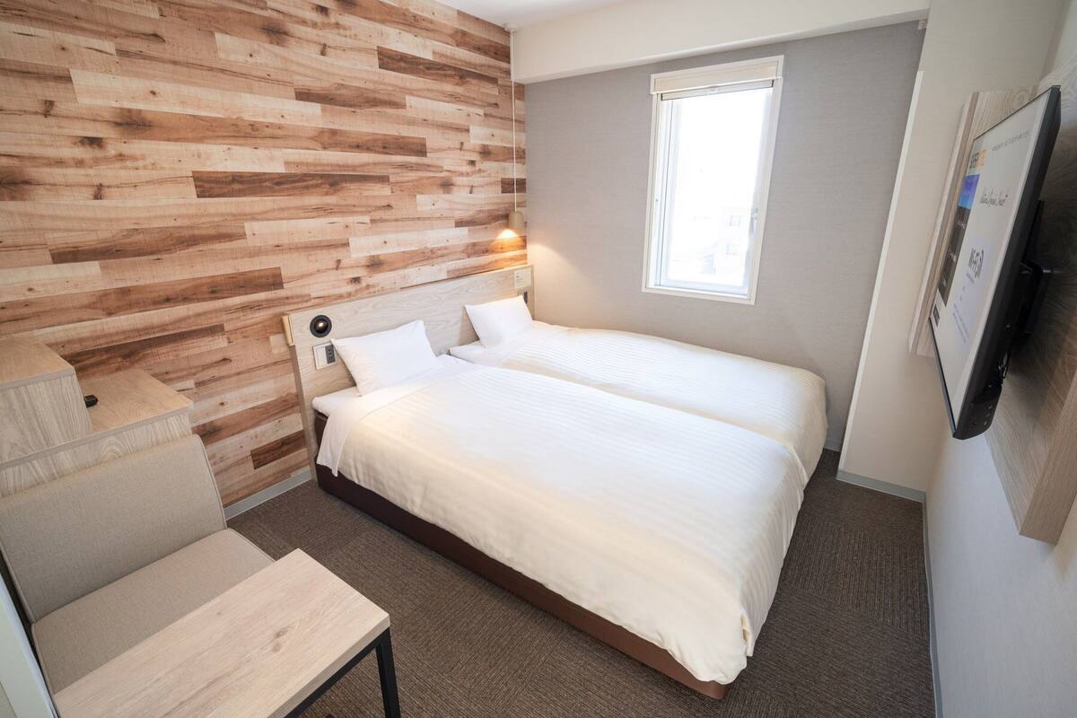 Habitación de hotel con pared de madera, cama con ropa blanca, una mesita y ventana con vistas.