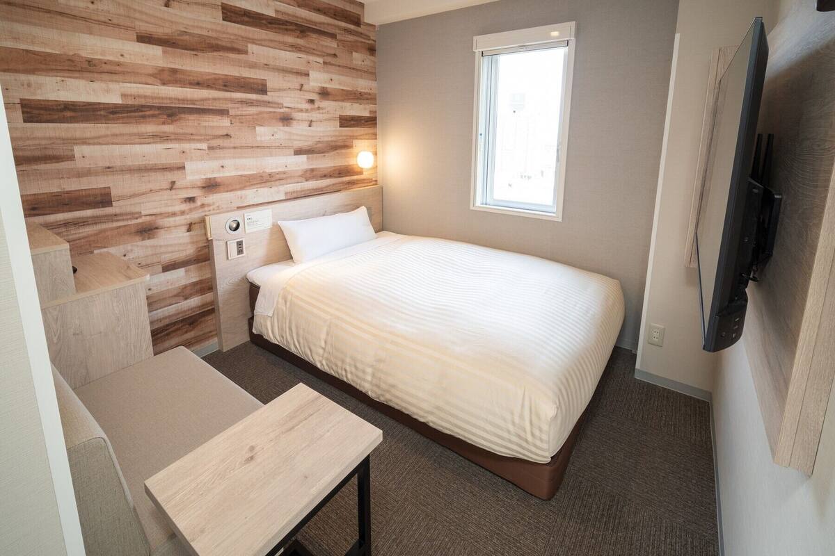 Un dormitorio pequeño y moderno con una pared de acento de madera, una cama individual, una mesita de noche, una ventana y un televisor de pantalla plana montado en la pared.