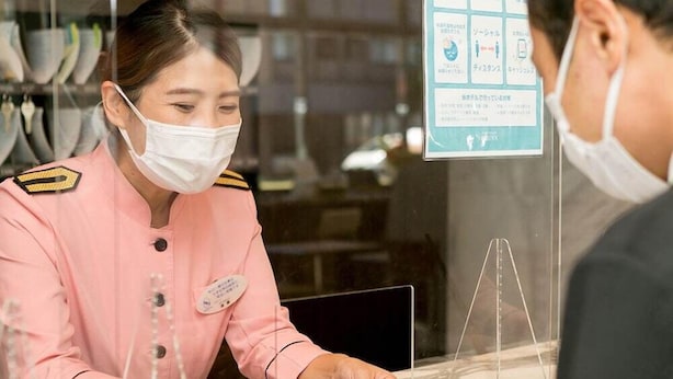 A hotel reception area with a staff member in a pink uniform behind the counter, a customer interacting with the staff, and a menu board visible on the wall. - Toyoko Inn Hakodate Ekimae Asaichi (HOKKAIDO)