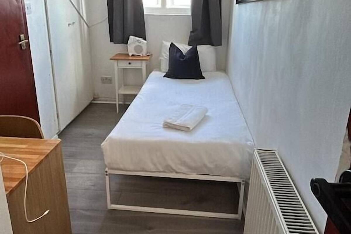 Une petite pièce, de taille modeste, comprenant un lit, un bureau, un radiateur et une fenêtre avec des rideaux.