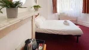 Una camera d'hotel con un letto, un tavolino con un bollitore e riviste, una pianta in vaso e una finestra con tende.