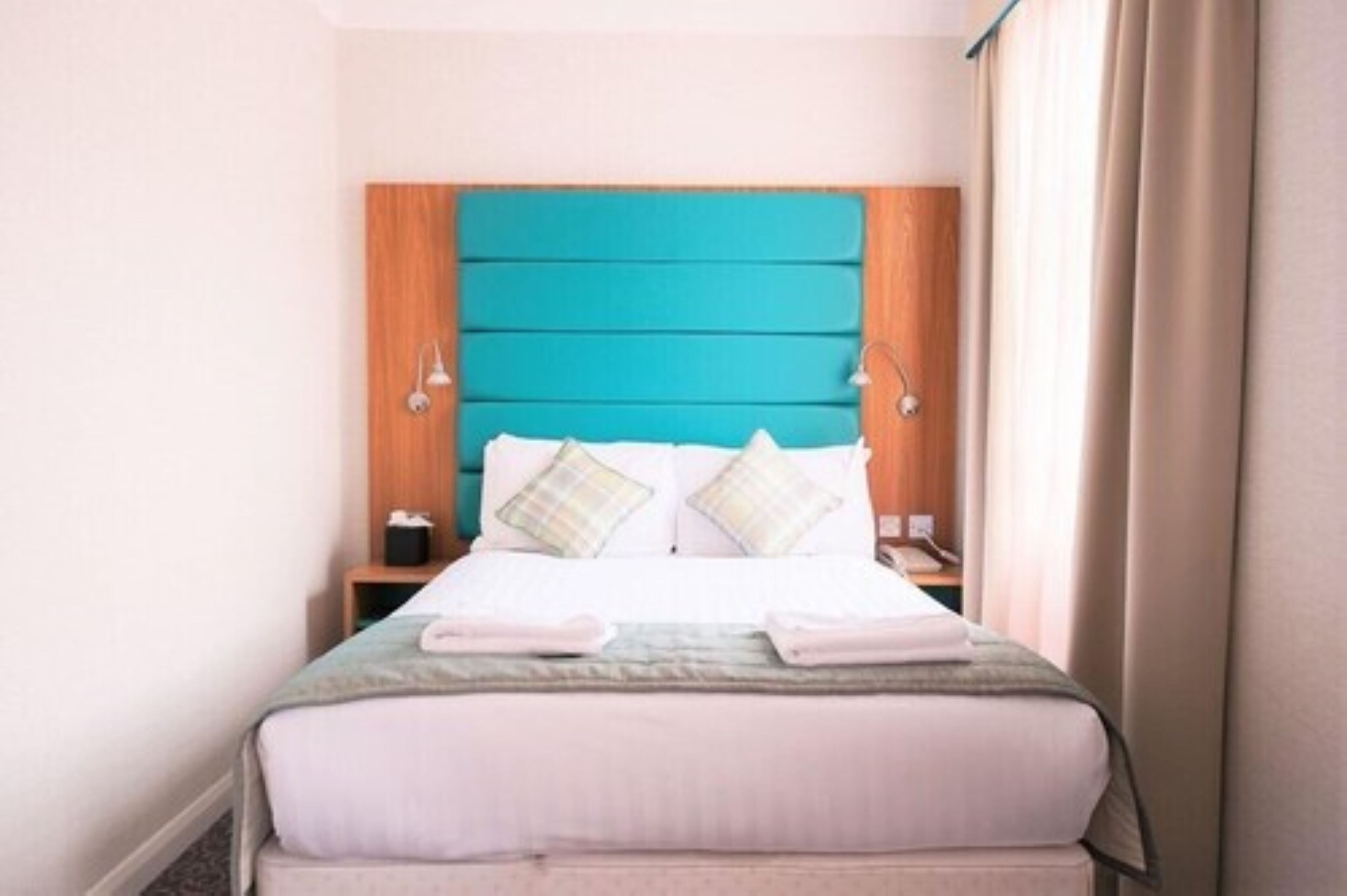 A neatly made bed with a teal headboard, two bedside tables, and a window with curtains.