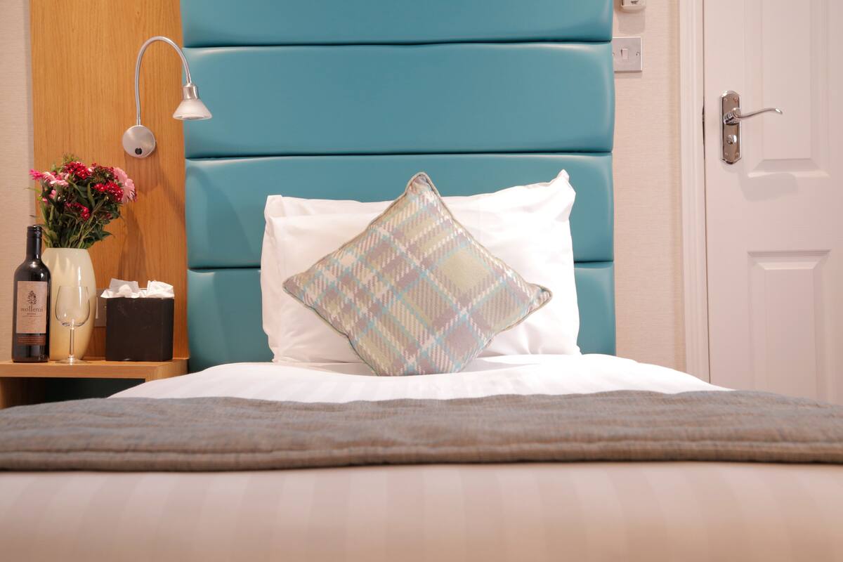 A neatly made bed with a patterned pillow, a bedside table with a bottle and glass, and a wall-mounted lamp.