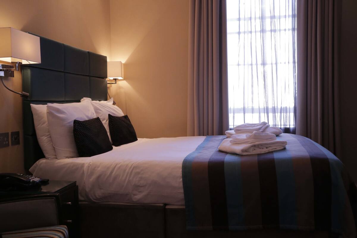 A hotel room with a large bed, a bedside table with a lamp, and a window with curtains.