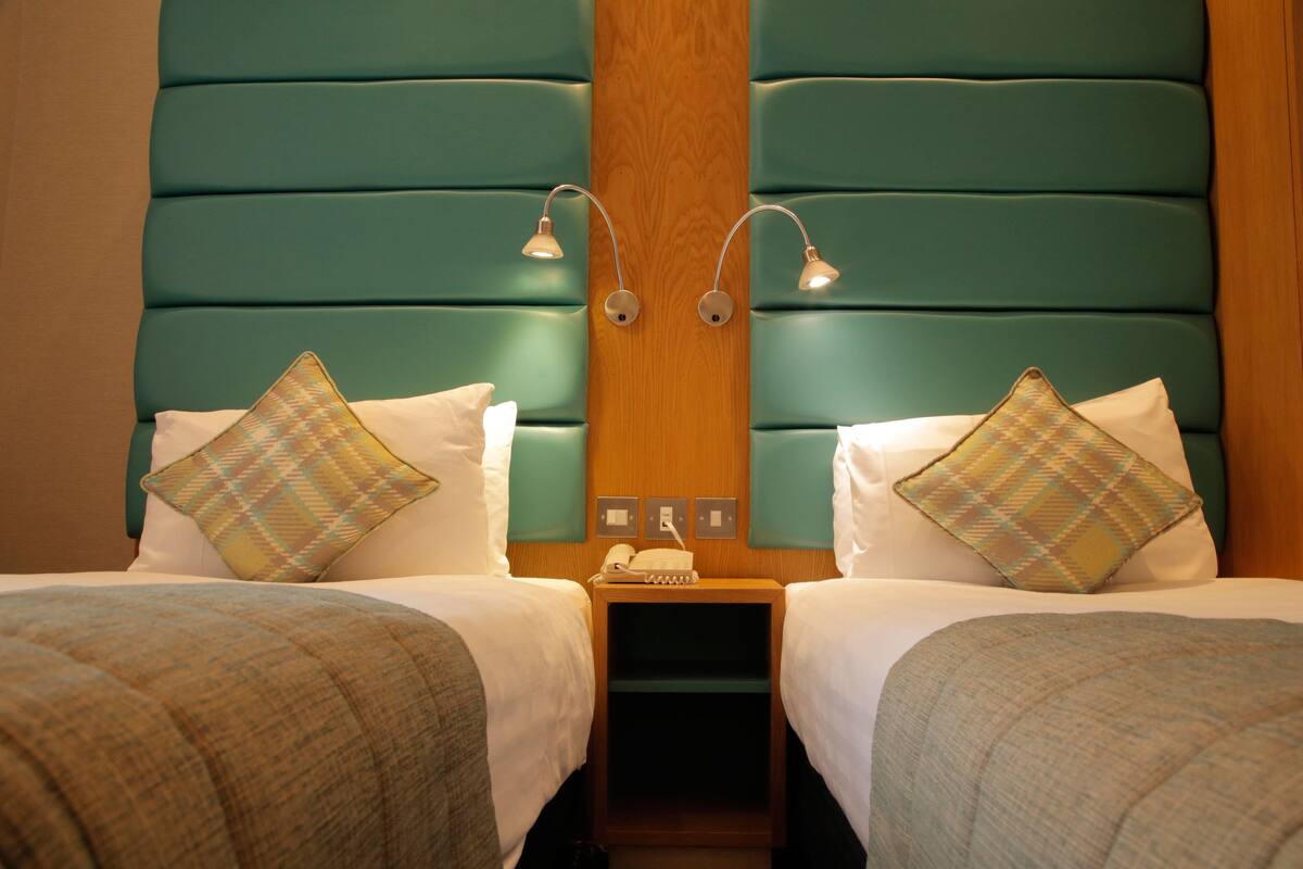 A hotel room with two beds, teal headboard, bedside lamps, and patterned pillows.