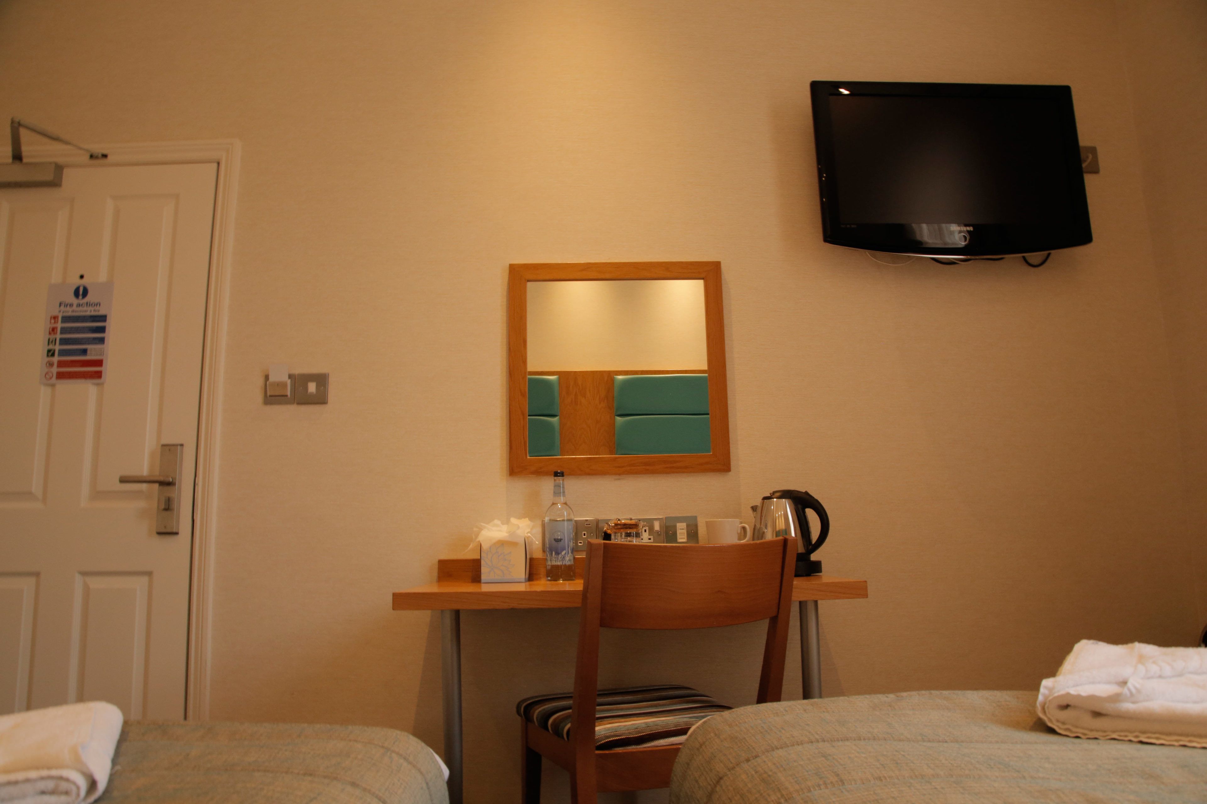 A hotel room with a bed, a desk with a mirror, a television, and a door with a fire action sign.