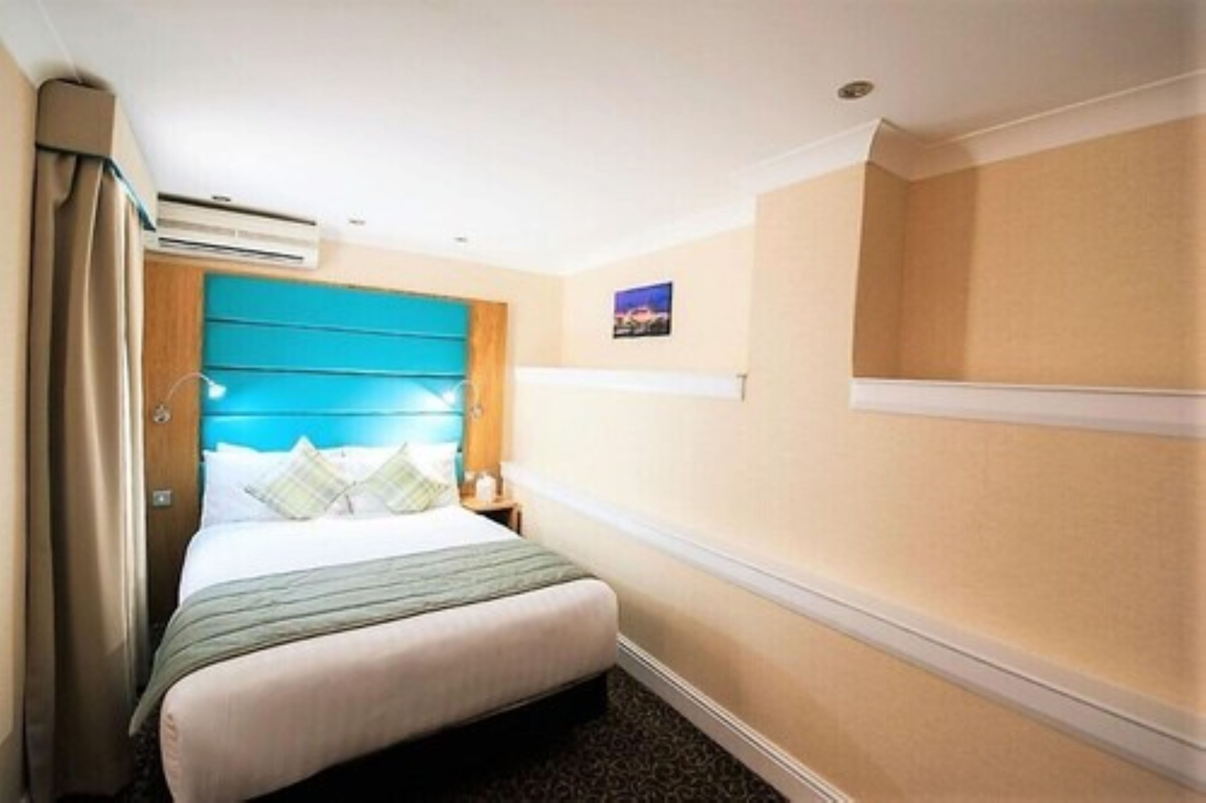 Habitación de hotel con cama, cabecera, mesita de noche, aire acondicionado de pared e imagen colgada en la pared.