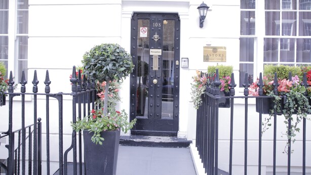 A black reception door with a sign above it, a white building facade, and a black iron fence with potted plants. - Astors Belgravia (London)