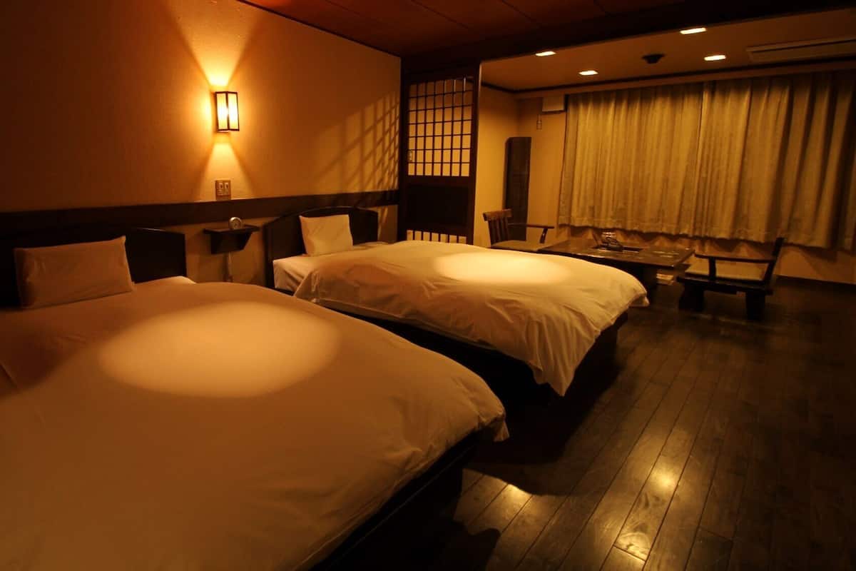 Habitación de hotel con dos camas, una mesita de noche, una lámpara, una silla y un ventanal con cortinas.