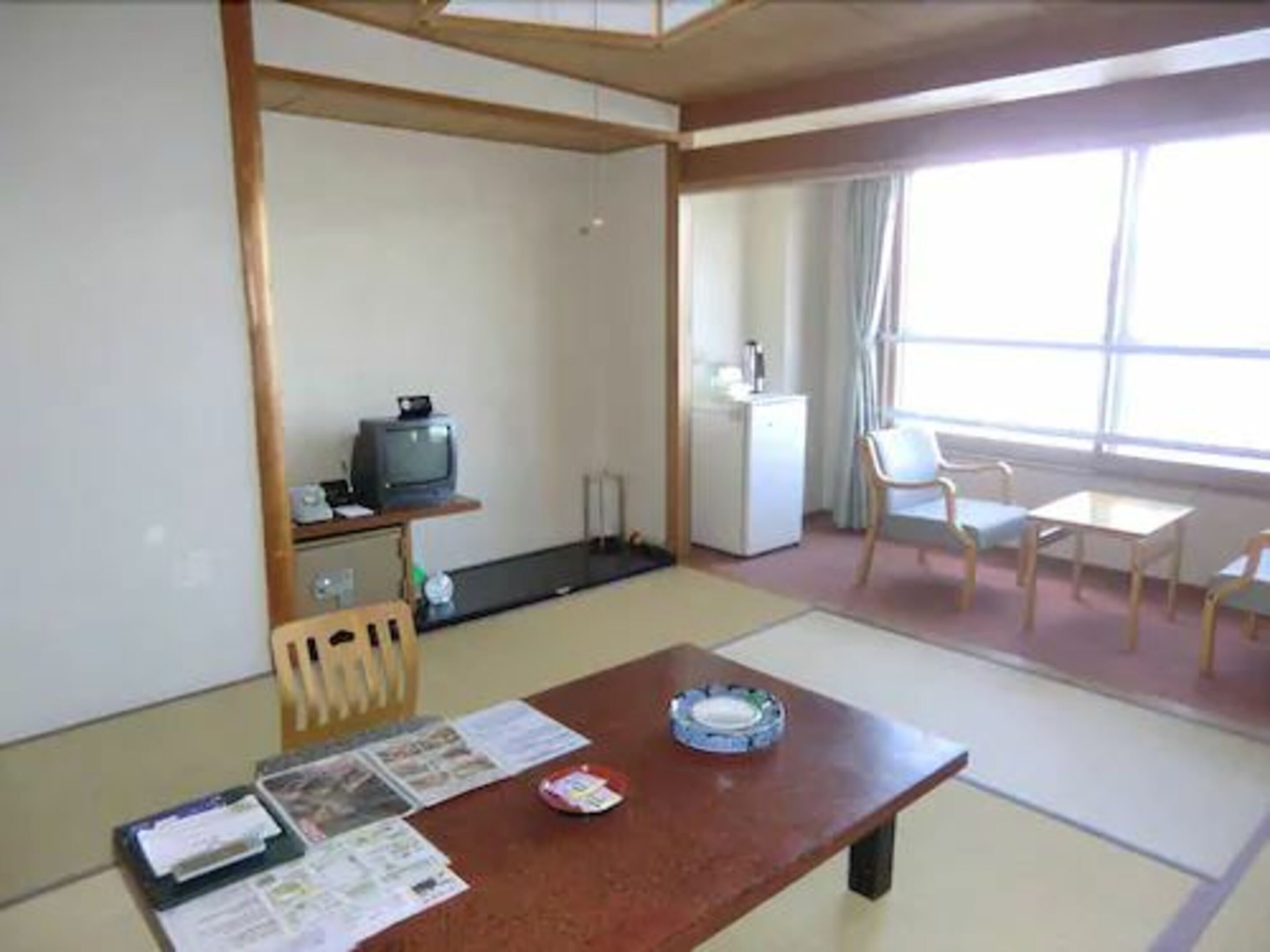 A hotel room with a wooden table, a television, a refrigerator, and a window with curtains.