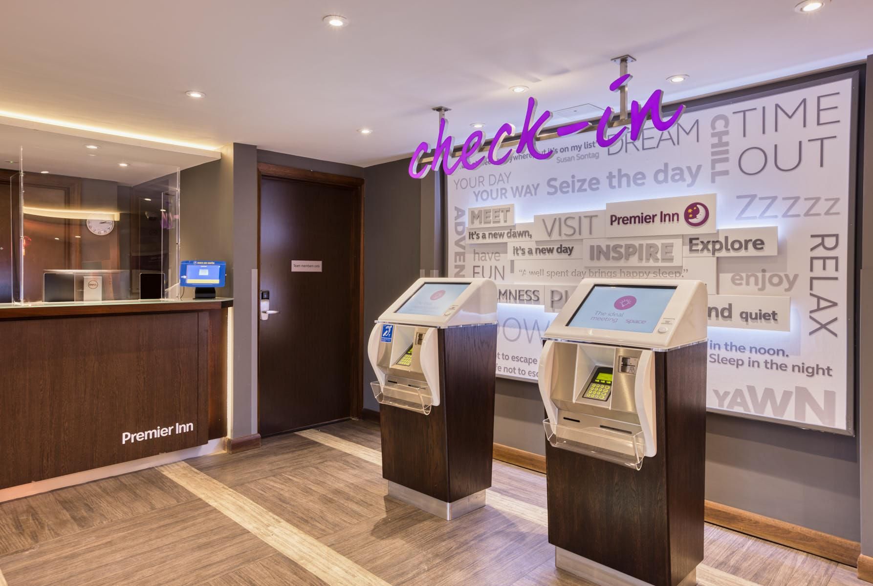 A hotel reception area with a digital kiosk, a reception desk, and a wall with motivational phrases.