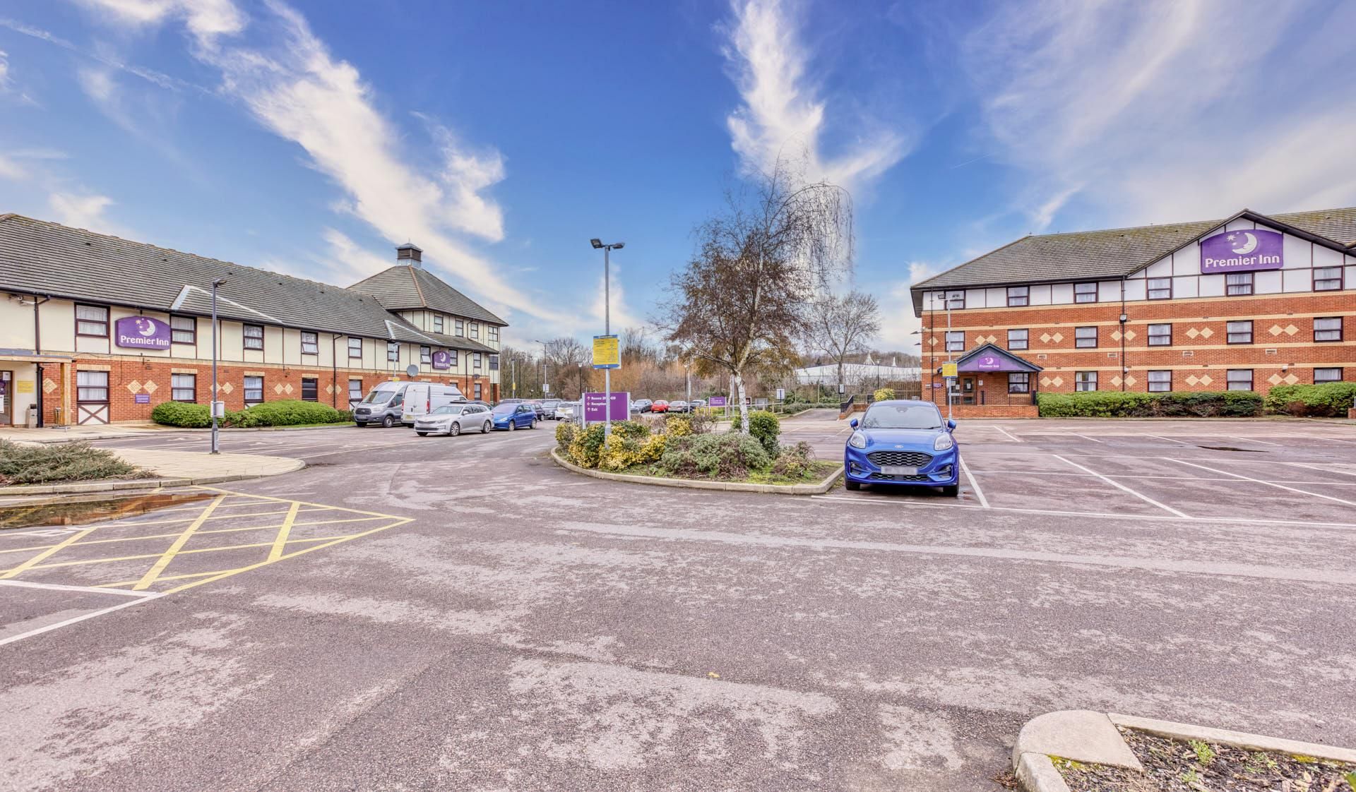 A Premier Inn hotel with a parking lot, a blue car in the foreground, and a clear sky.