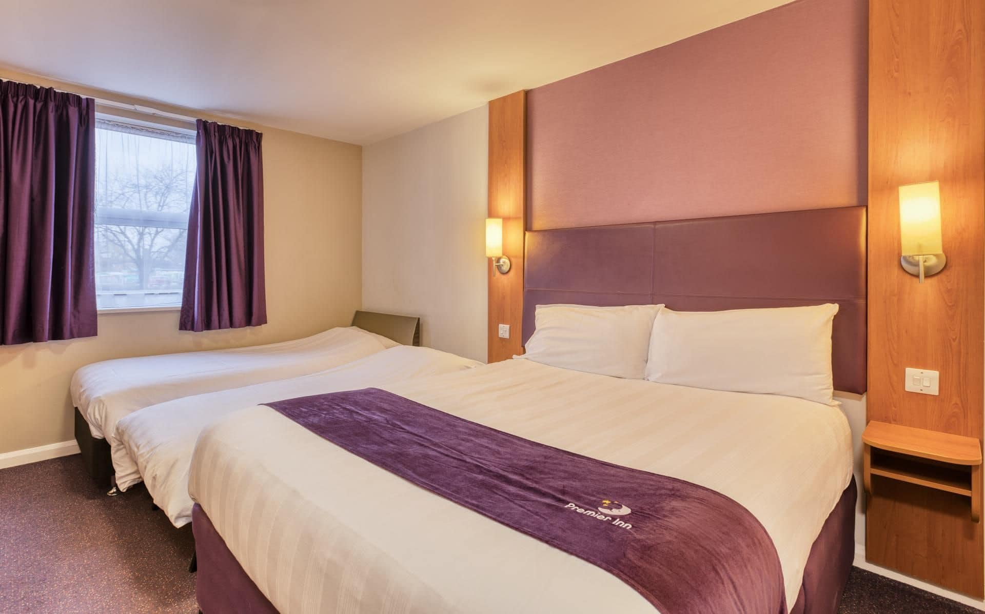 A hotel room with two beds, a headboard, bedside lamps, and a window with curtains.