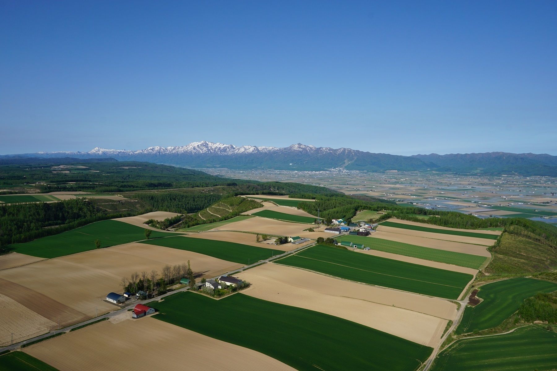 Une vue aérienne d’un paysage rural, avec des champs, un petit groupe de bâtiments et, au loin, des montagnes enneigées.