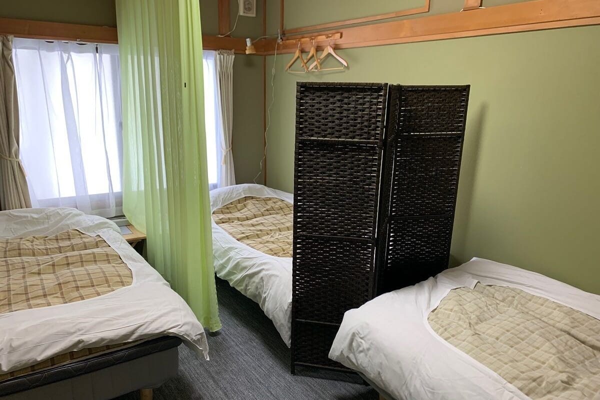 Une pièce avec trois lits, chacun recouvert d’un drap blanc et d’une couverture à carreaux. Une fenêtre est ornée d’un rideau vert, et un paravent noir tissé sépare les lits.