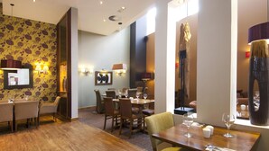 Un espace repas avec des tables et des chaises en bois, des lampes fixées au mur et un papier peint à motifs.