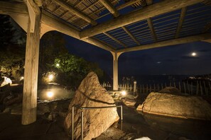 Um pavilhão ao ar livre com estrutura de telhado de madeira, rodeado por pedras grandes e um corpo d'água.