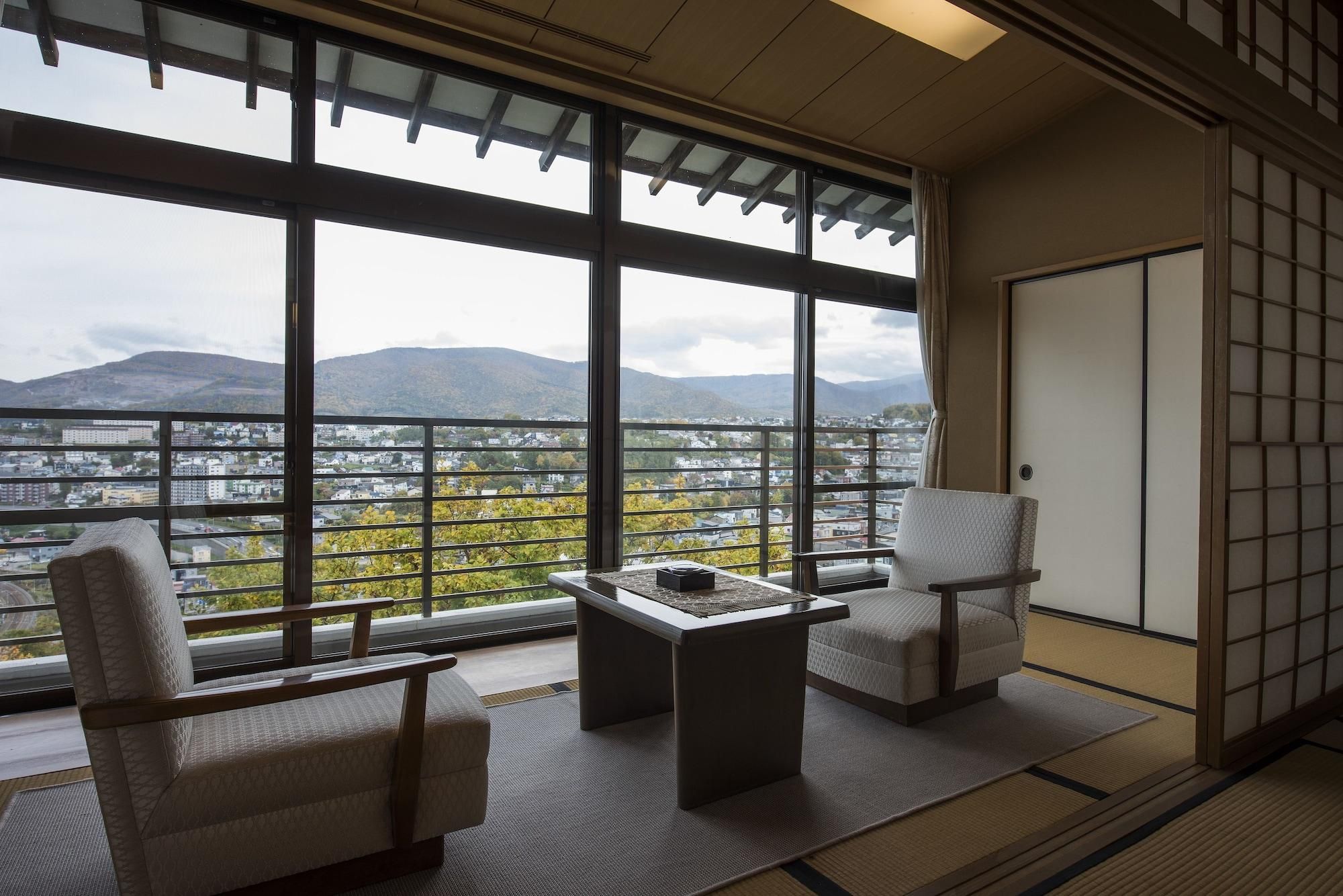 Quarto com vista para a paisagem urbana, duas cadeiras, uma mesa pequena e piso tradicional de tatame.