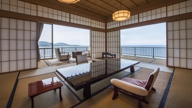 A traditional Japanese room with tatami flooring, a low table, and seating. Large windows offer a view of the sea and distant hills.