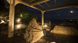A covered outdoor area with wooden beams and a metal railing, surrounded by large rocks and a view of the water.