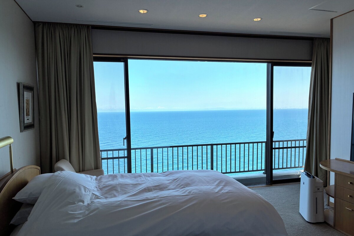 A hotel room with a large bed, a desk, a chair, a waste bin, and a view of the sea.