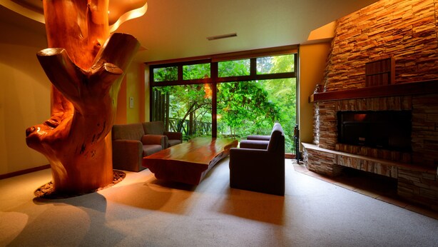 A modern living room with a large wooden sculpture, a stone fireplace, and a view of lush greenery through large windows. - Hotel Daiheigen (Otofuke)