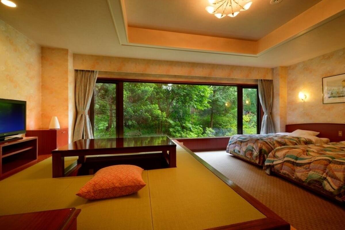 A hotel room with a bed, a television, a seating area with cushions, and a large window offering a view of the outdoors.