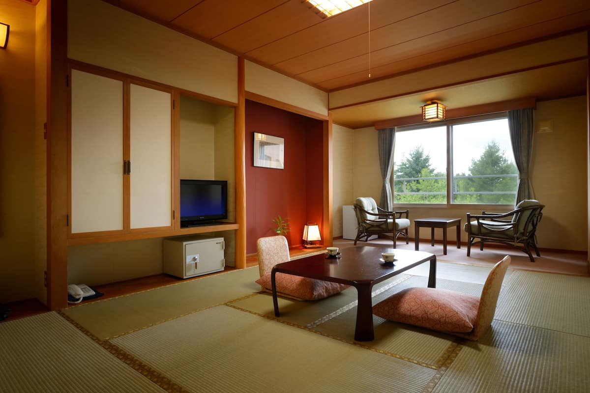 A traditional Japanese-style room with tatami flooring, a low table with cushions, a television, and a view of greenery outside.