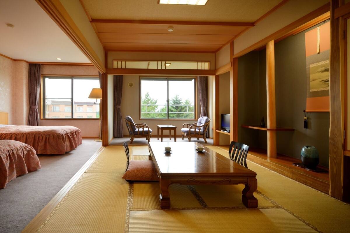 A hotel room with two beds, a dining table, chairs, a TV, and large windows.