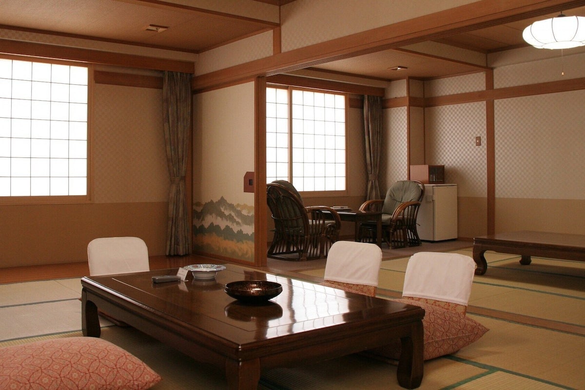 A traditional Japanese room with tatami flooring, a low table, chairs, and a view of the outside through shoji screens.