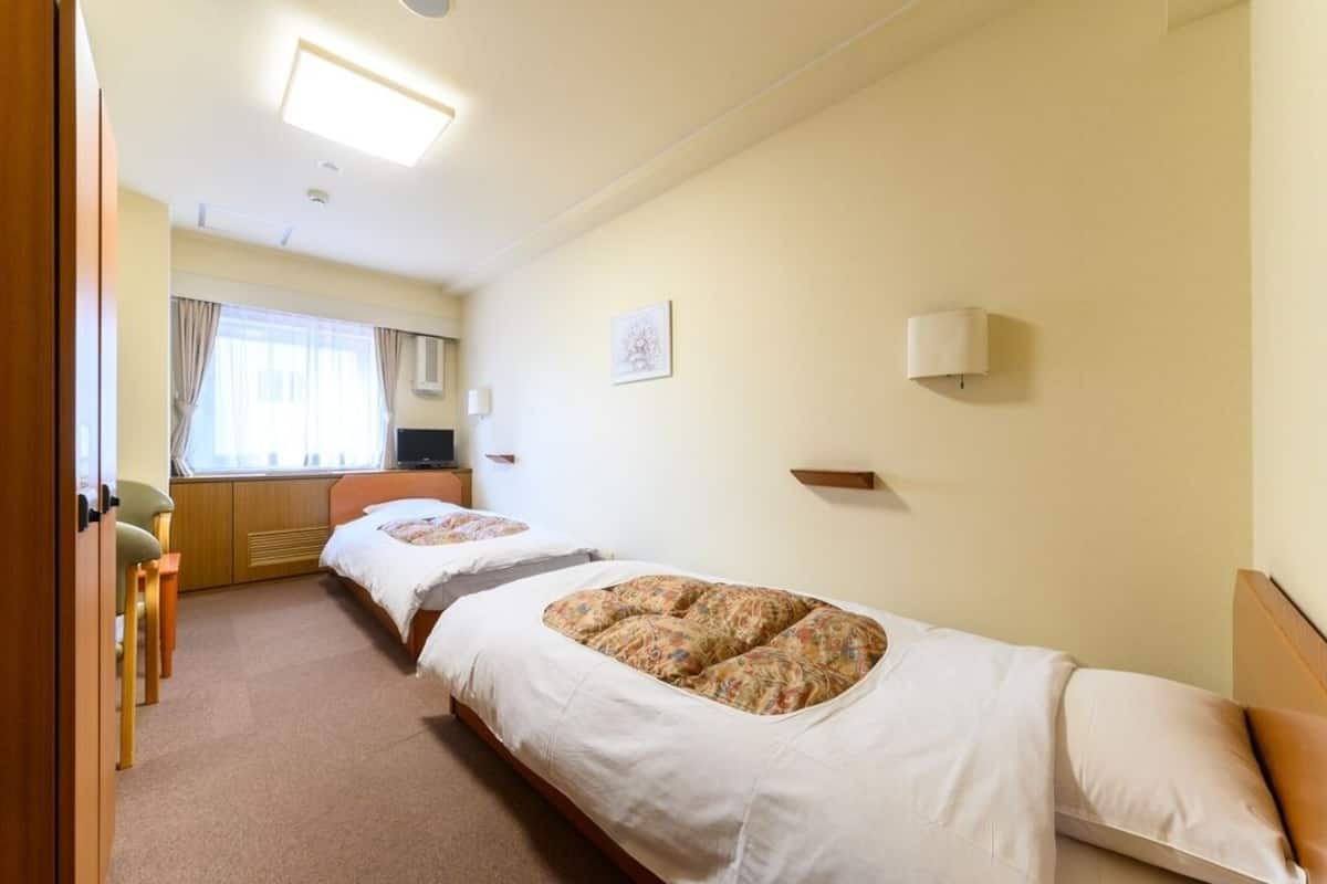 A hotel room with two beds, a desk, a chair, a window with curtains, and a wall-mounted lamp.
