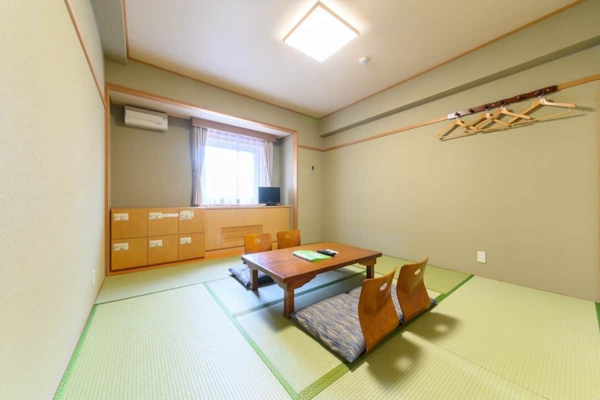 A room with tatami flooring, a wooden table, a sofa, a window with curtains, a wall-mounted air conditioner, and a clothes drying rack.