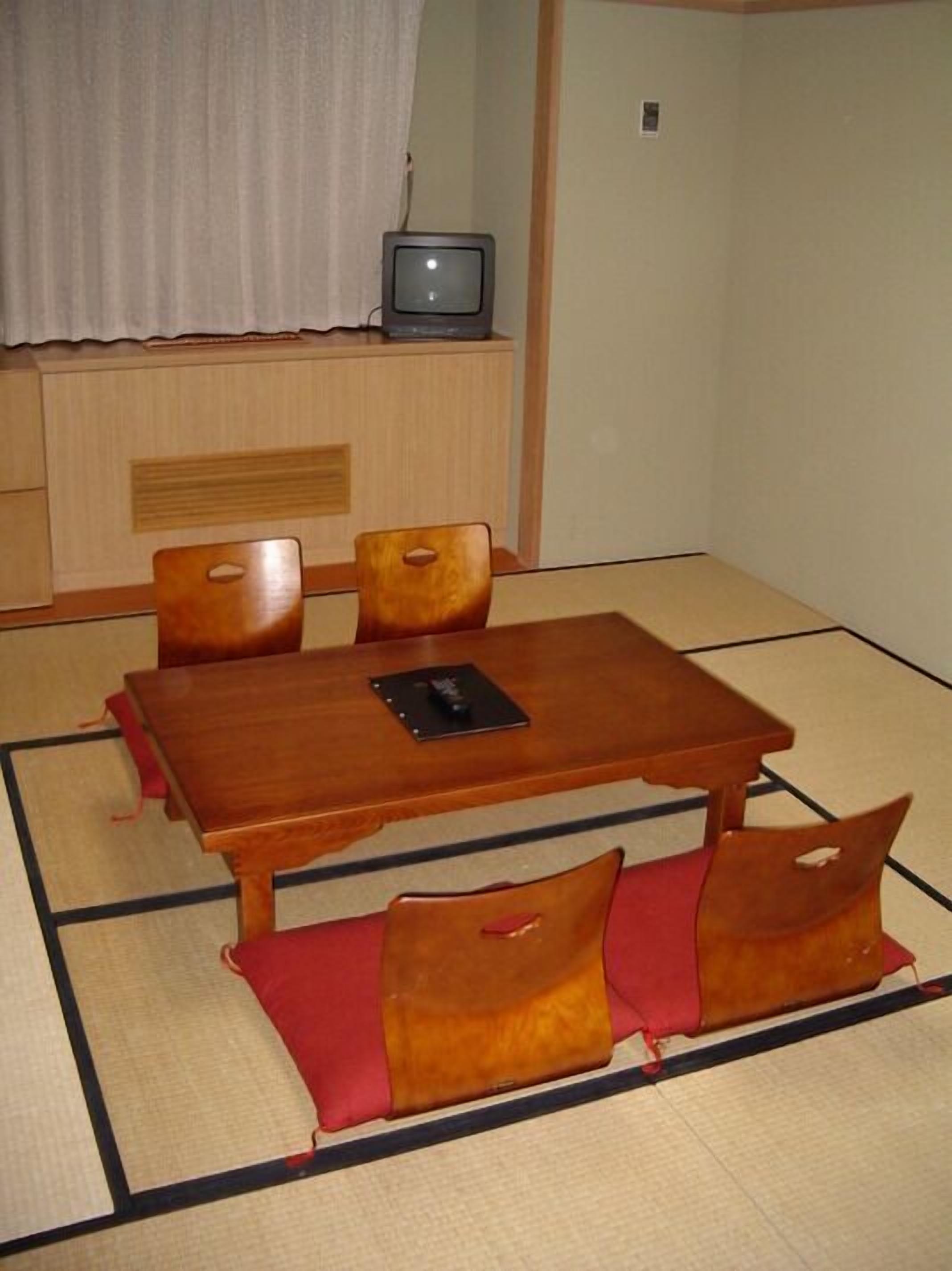 Een kamer met tatami matten, een houten tafel, vier stoelen en een televisie op een plank.