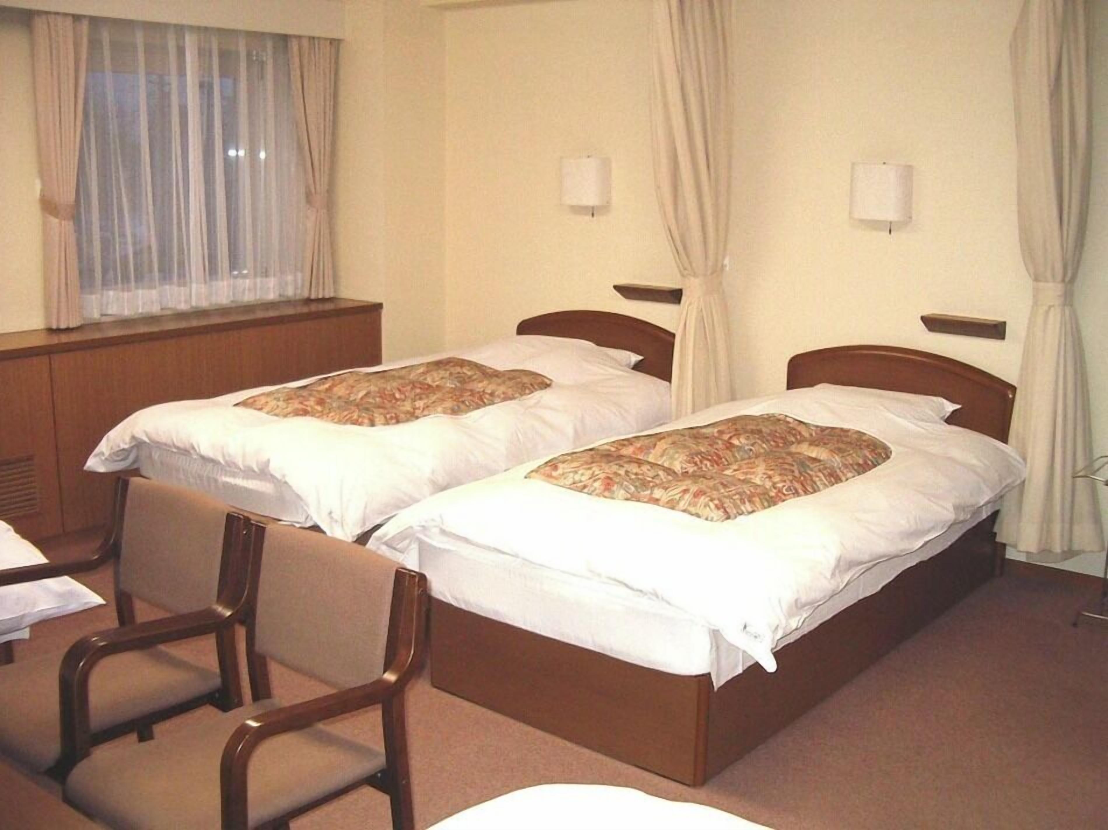 Een hotelkamer met twee eenpersoonsbedden, een houten hoofdbord, een tafeltje en een stoel.