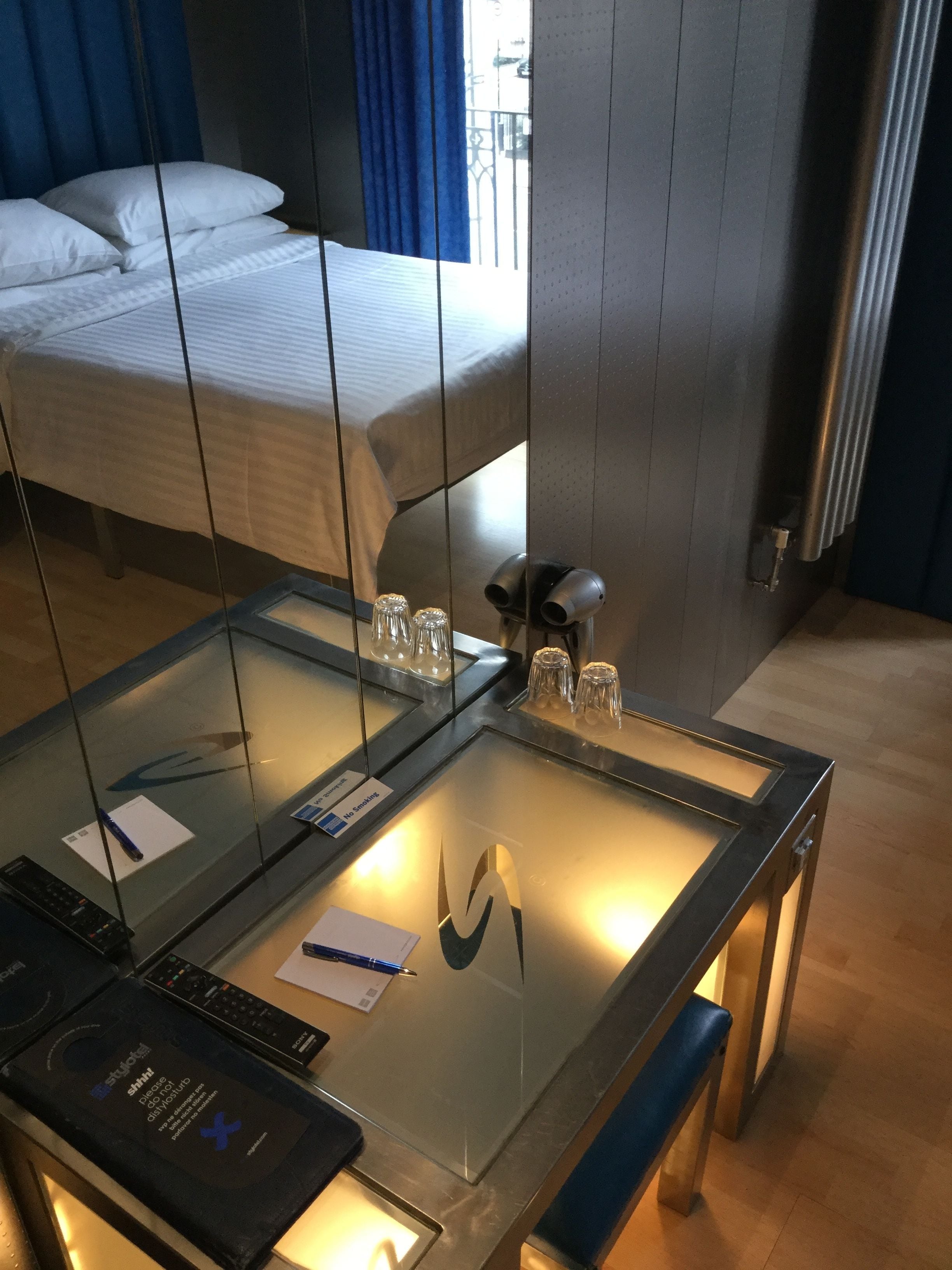 Ein Hotelzimmer mit einem Bett, einem Glastisch mit einem Laptop und einem Spiegel, der das Bett reflektiert.