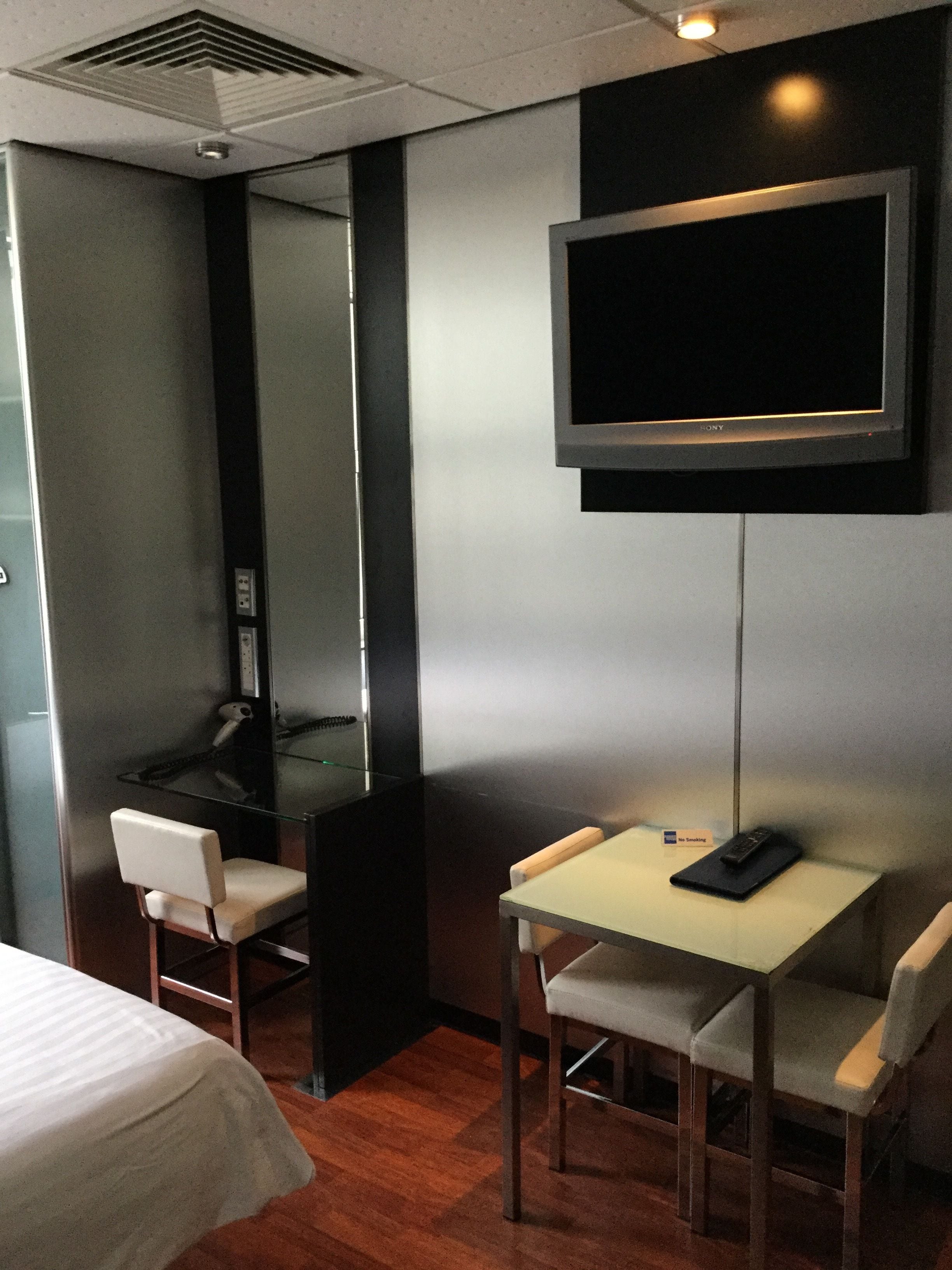 A hotel room with a bed, a desk with a chair, a television mounted on the wall, and a small table with a chair.