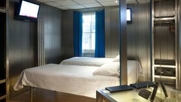 Une chambre d’hôtel avec deux lits, un téléviseur fixé au mur, une fenêtre avec des rideaux bleus et un bureau avec un ordinateur et des lunettes.