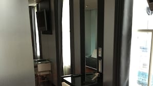 A hotel room with a wooden floor, a desk with a chair, a mirror, and a window with curtains.