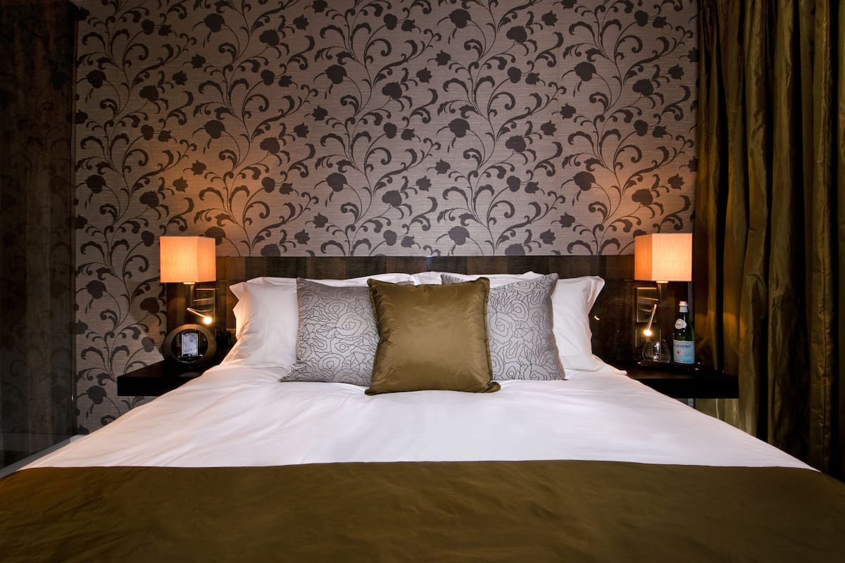 A neatly made bed with white linens and a brown throw blanket, flanked by two bedside tables with lamps and a bottle of water.