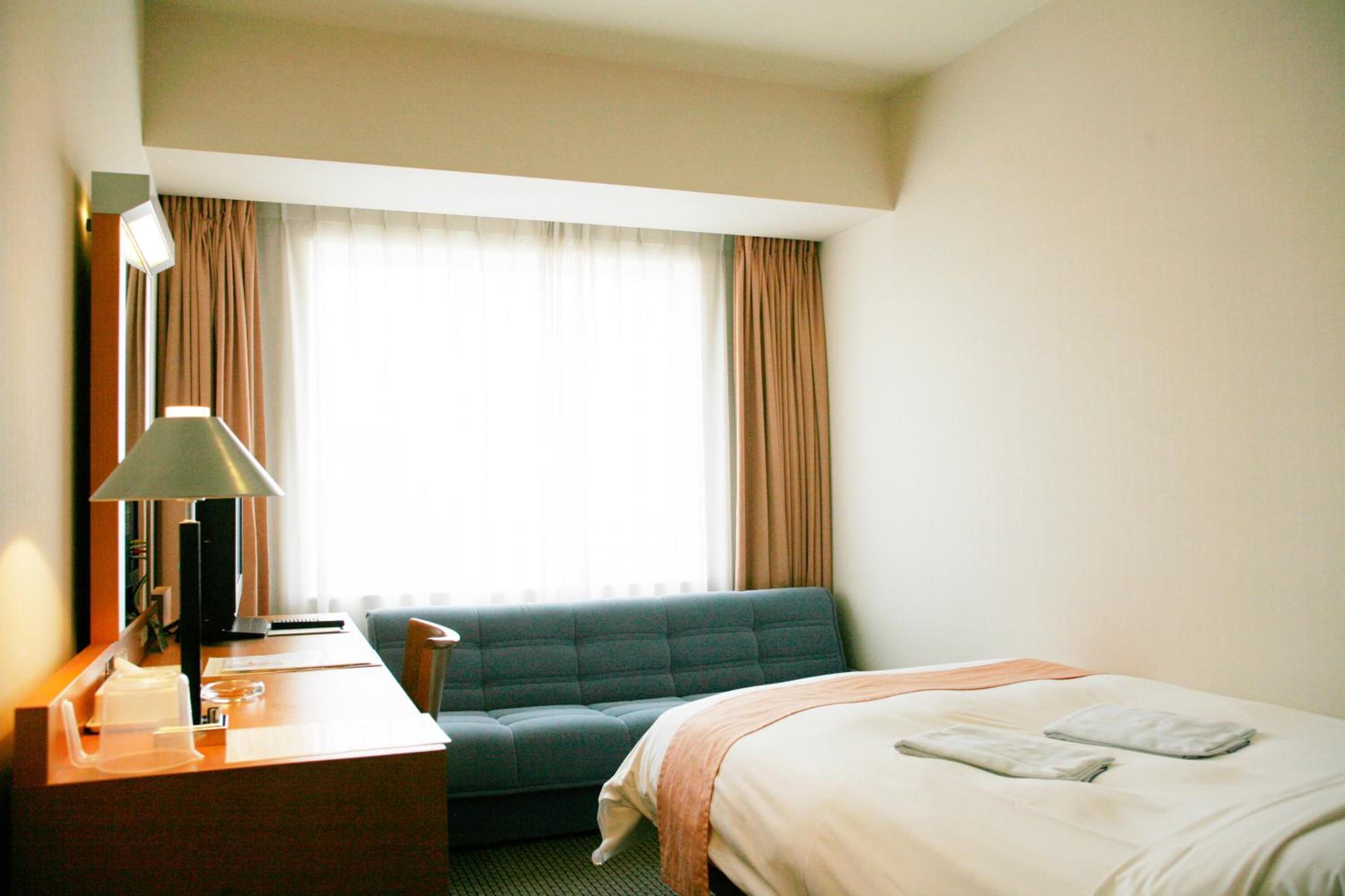 Une chambre d’hôtel avec un lit, un bureau avec une lampe, un canapé et une fenêtre avec des rideaux.