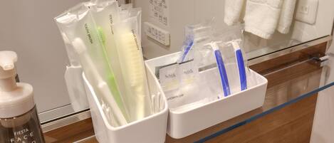 Una encimera de baño con artículos de aseo, incluyendo una botella de espuma limpiadora para cara y manos, un cepillo de dientes verde, uno blanco y uno azul dentro de un gorro de ducha.