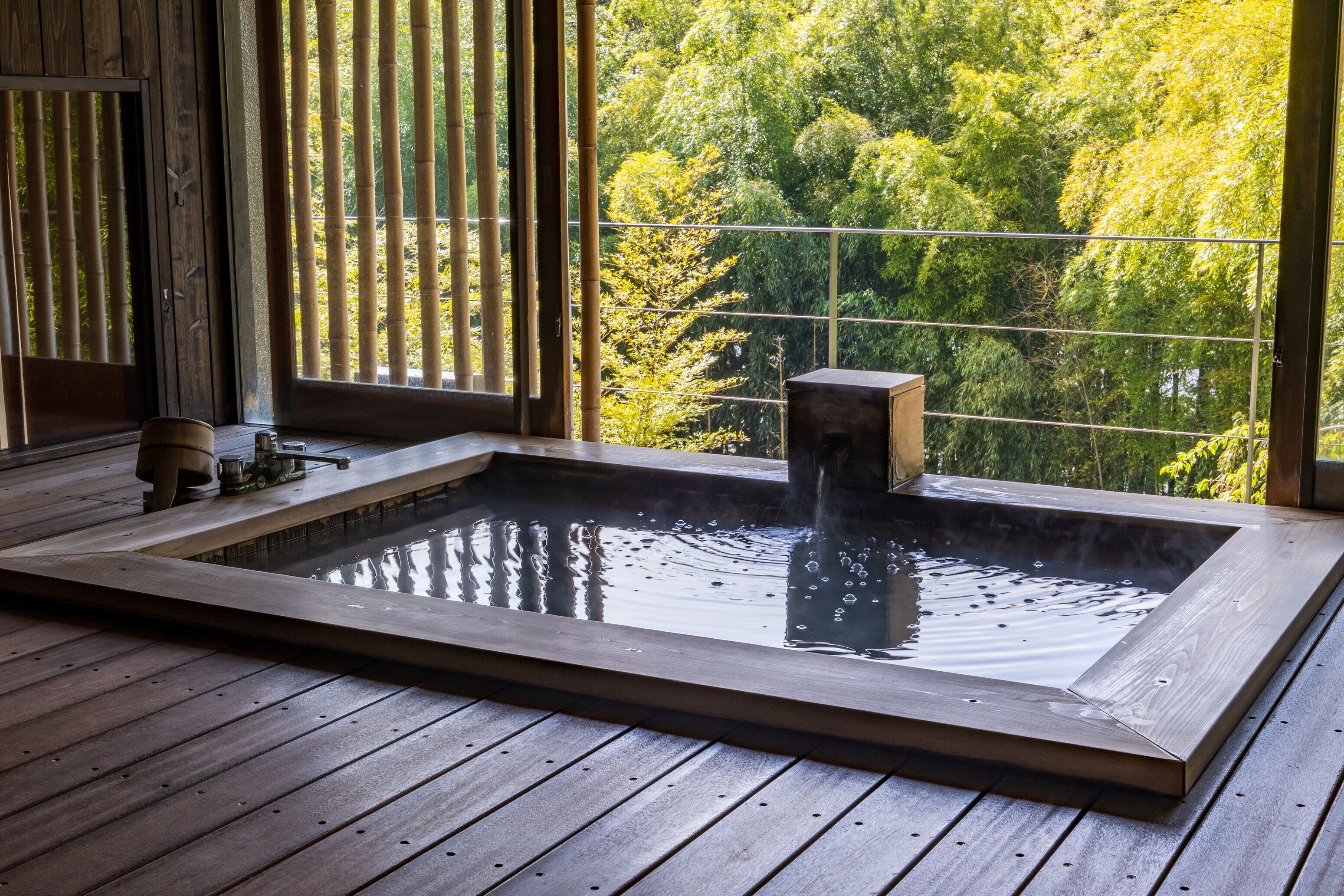 A traditional Japanese onsen with a wooden deck, a hot spring bath, and a view of lush greenery through bamboo blinds.